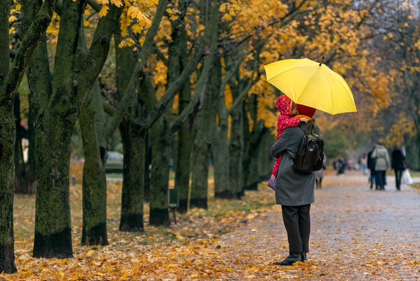 Mamá sostiene a un niño pequeño en sus brazos mientras está de pie bajo un paraguas amarillo en un parque de otoño sobre un fondo de follaje amarillo. - Foto, Imagen
