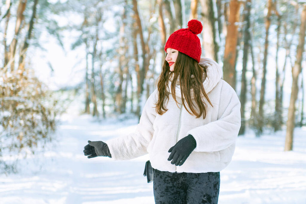 Szczęśliwa młoda kobieta w czerwonym kapeluszu z zamkniętymi oczami ciesząca się chwilą w śnieżnym lesie. Słoneczny dzień w zimowym parku - Zdjęcie, obraz