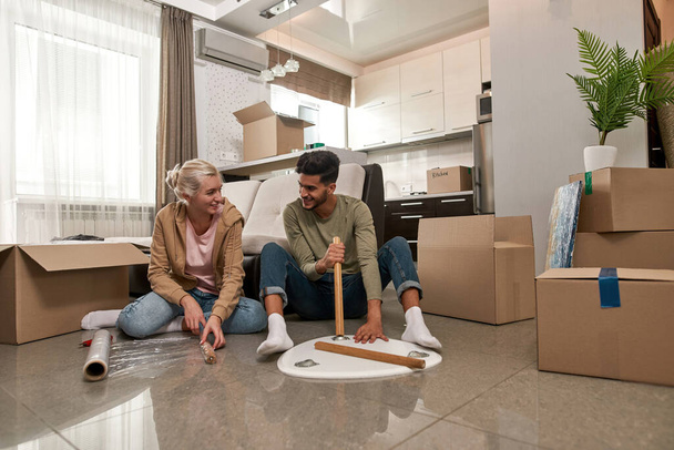 Caucasian girl helping her middle eastern husband or boyfriend packing table on floor. Young smiling multiracial couple looking at each other and moving to their new apartment. Home relocation - Photo, Image