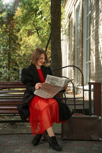 Cheerful young woman in red dress and black coat sits on bench reading newspaper and laughing - Foto, Imagen