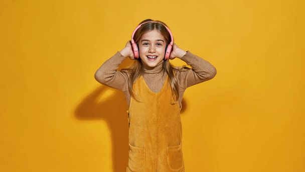Vista frontal de una niña caucásica sonriente escuchando música en auriculares. Hija de la generación de zoómeros. Concepto de estilo de vida infantil moderno. Aislado sobre fondo amarillo en estudio. Copiar espacio - Foto, imagen