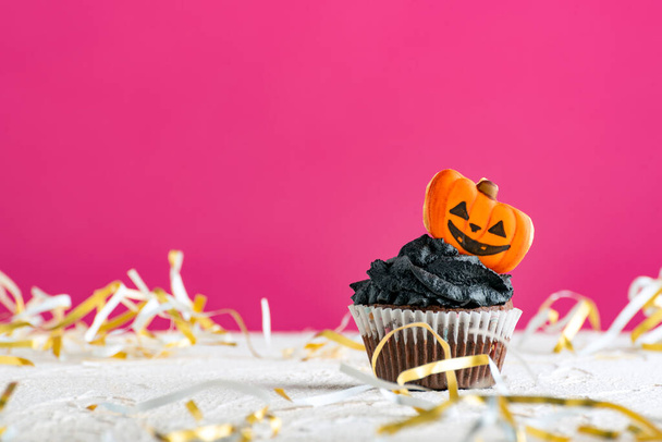 Cupcake decorated with black cream and gingerbread cookies pumpkins on pink background. Festive Halloween muffin. - Photo, Image