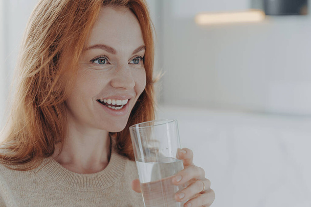 Close up shot of ginger woman drinks pure water from glass enjoys refreshing drink healthy liquid smiles broadly focused into distance wears jumper poses indoor blank space follows healthy lifestyle - Photo, Image