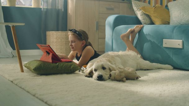 Young girl lying on mild carpet, choosing video on digital tablet computer, dog feeling tired and trying to sleep, spending leisure time at home with pet. Golden retriever. - Fotoğraf, Görsel