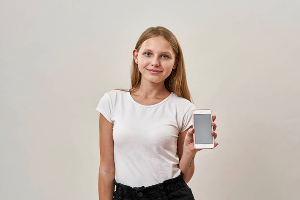 Smiling caucasian teenage girl showing mobile phone and looking at camera. Blonde female child of zoomer generation. Modern youngster lifestyle. Isolated on white background. Studio shoot. Copy space - Photo, Image