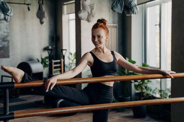 Izgatott, karcsú, vörös hajú nő áll a modern fitness stúdióban a kezével és fél lábával a balettkorláton, nyújtózkodik és kamerába néz, kényelmes fekete sportruházatot visel. - Fotó, kép