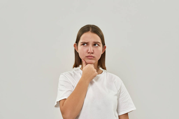 Caucasian teenage girl thinking about something. Cute female of zoomer generation wearing t-shirt. Concept of modern youngster lifestyle. Isolated on white background. Studio shoot. Copy space - Φωτογραφία, εικόνα