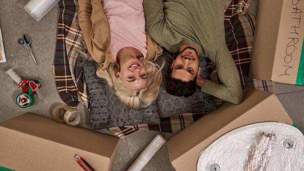 Top view of young smiling multiracial couple lay and look at camera on blanket on floor with boxes and things for packing. European girl and middle eastern man moving to new apartment. Home relocation - Photo, image
