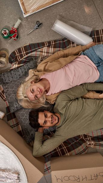 Top view of young smiling multiethnic couple lying and looking at camera on blanket on floor with cardboard boxes and things for packing. Blonde girl and man moving to new apartment. Home relocation - Foto, afbeelding