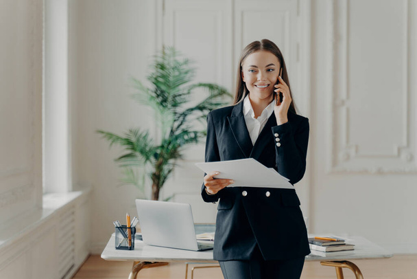 Профессиональная молодая деловая леди в формальном костюме смотрит в сторону, разговаривая по телефону и держа в руках записки, бизнес-лидер смеется, когда слышит хорошие новости, счастливая работница офиса, стоящая в офисе - Фото, изображение