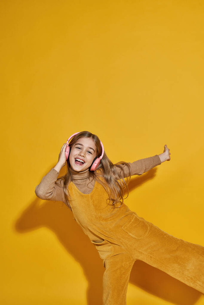 Joyful european little girl listening music in headphones and dancing. Female child of zoomer generation. Concept of modern childhood lifestyle. Isolated on yellow background. Studio shoot. Copy space - Foto, Bild