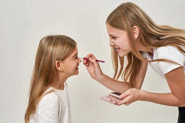 Partial side of teenage girl painting glitter eyelid of little sister with cosmetic brush. Caucasian sisters of zoomer generation. Female beauty. Family caring. Isolated on white background in studio - Foto, Bild