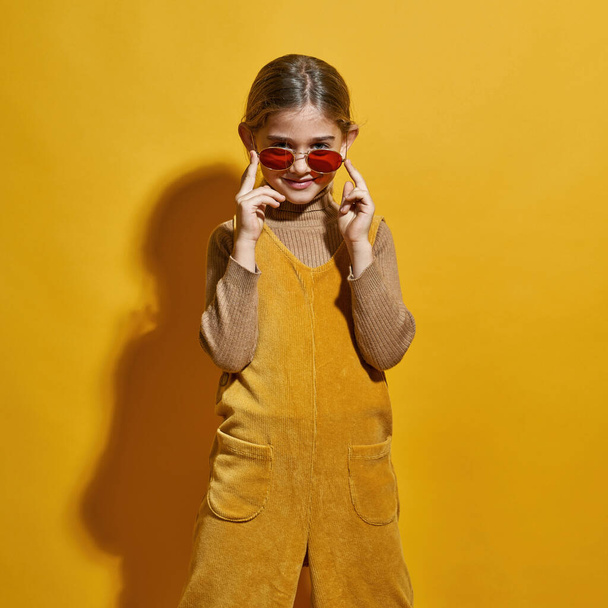Front view of caucasian little girl wearing glasses looking at camera. Female child of zoomer generation. Concept of modern childhood lifestyle. Isolated on yellow background. Studio shoot. Copy space - Photo, Image