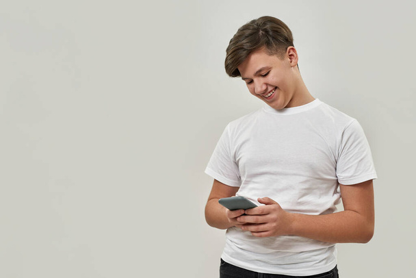 Smiling caucasian teenage boy browsing his smartphone. Guy of zoomer generation wearing t-shirt. Concept of modern youngster lifestyle. Isolated on white background. Studio shoot. Copy space - Fotoğraf, Görsel