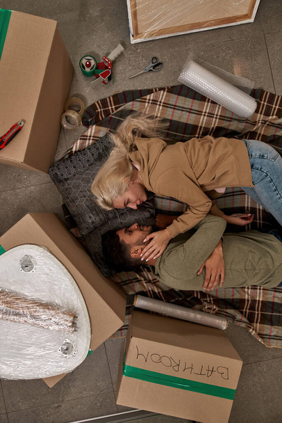 Top view of young multiracial couple lay and look at each other on blanket on floor with boxes and things for packing. Girl touch face of boyfriend. Moving to new apartment. Relationship and closeness - Photo, image