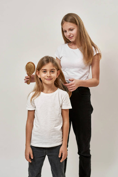 Teenage girl combing little sister hair with comb. Blonde smiling caucasian sisters of zoomer generation. Concept of family caring and relationship. Isolated on white background in studio. Copy space - Photo, image