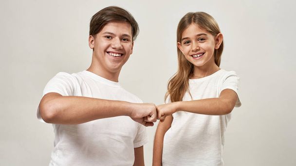 Teenage boy and little girl bumping fists of each other. Smiling caucasian brother and sister of zoomer generation looking at camera. Modern youngster lifestyle. Isolated on white background in studio - Fotó, kép