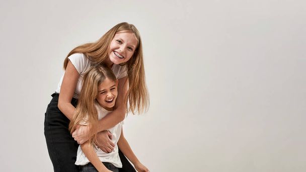 Teenage girl hugging her little sister and looking at camera. Cheerful caucasian sisters of zoomer generation enjoying time together. Modern youngster lifestyle. White background in studio. Copy space - Zdjęcie, obraz