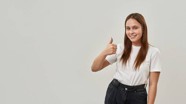 Smiling caucasian female teenage showing thumb up and looking at camera. Cute girl of zoomer generation. Concept of modern youngster lifestyle. Isolated on white background. Studio shoot. Copy space - Zdjęcie, obraz