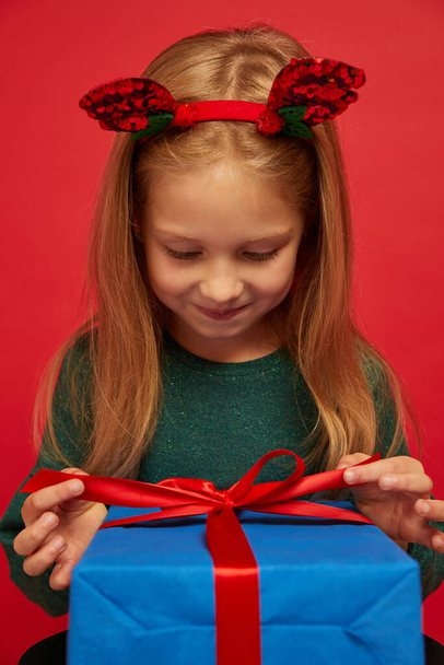 Smiling funny child (kid, girl) in Reindeer Hairband. Holding Christmas gift in hand. Christmas concept. Shooting on red background - Valokuva, kuva