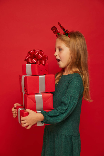 Smiling funny child (kid, girl) in Reindeer Hairband. Holding Christmas gifts in hand. Christmas concept. Shooting on red background - Foto, imagen