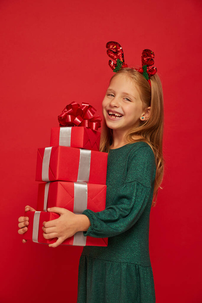 Smiling funny child (kid, girl) in Reindeer Hairband. Holding Christmas gifts in hand. Christmas concept. Shooting on red background - Foto, afbeelding