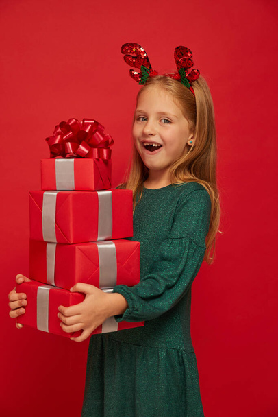 Smiling funny child (kid, girl) in Reindeer Hairband. Holding Christmas gifts in hand. Christmas concept. Shooting on red background - Zdjęcie, obraz