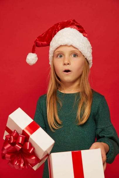 Smiling funny child (kid, girl) in Santa red hat opening  Christmas gift box in hand. Christmas concept. Shooting on red backgroun - Foto, afbeelding