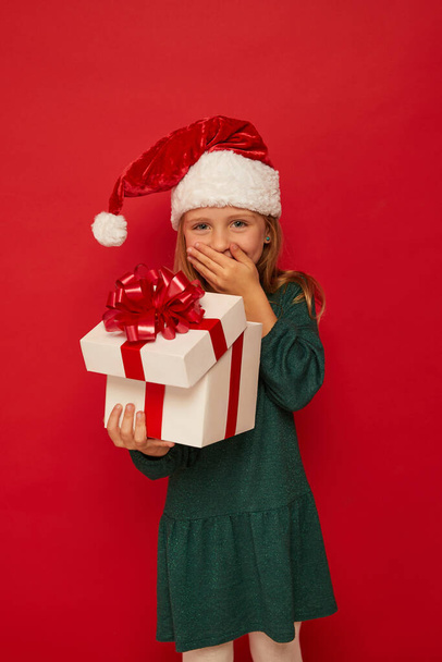 Smiling funny child (kid, girl) in Santa red hat opening  Christmas gift box in hand. Christmas concept. Shooting on red backgroun - Foto, Imagem