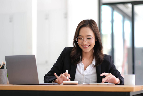 Прекрасна азіатська бізнесменка аналізує таблиці за допомогою калькулятора ноутбука в офісі... - Фото, зображення