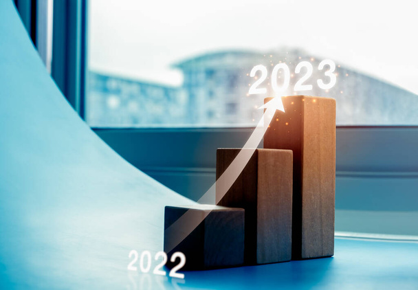 Λαμπερή άνοδο μέχρι βέλος στο ξύλο μπλοκ βήματα διάγραμμα ως γράφημα από το έτος 2022 έως 2023 σε μπλε φόντο με αστικό τοπίο, διαδικασία ανάπτυξης των επιχειρήσεων, το κέρδος, τον πλούτο, τις τάσεις, τις έννοιες οικονομικής βελτίωσης. - Φωτογραφία, εικόνα