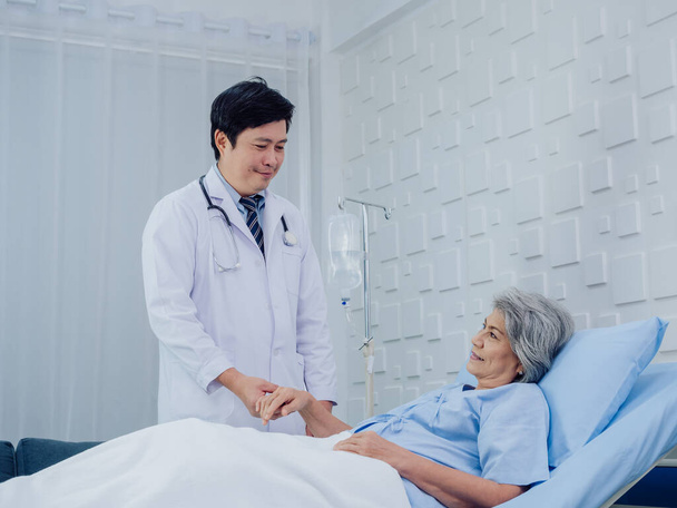 A kedves ázsiai férfi orvos fehér öltönyben meglátogatja, beszél és támogatást nyújt, kezében boldog idős nő beteg világoskék ruhában feküdt az ágyon sóoldatban a kórházi szobában. - Fotó, kép