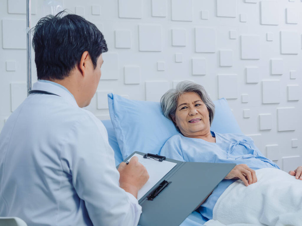Glücklich lächelnde asiatische Seniorin, alte Patientin, die mit Kochsalzlösung im Bett liegt und mit dem Arzt diskutiert, während er im Krankenhauszimmer Notizen macht. Gesundheitsversorgung und medizinisches Konzept. - Foto, Bild