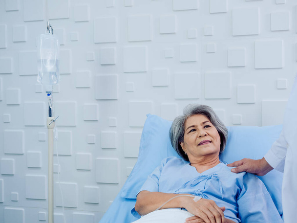 Glückliches Lächeln asiatische Senioren, ältere Patientin in hellblauem Kleid, die mit Kochsalzlösung im Bett liegt, während der Arzt in Weiß ihre Schulter berührt, spricht und berät und tröstet. - Foto, Bild