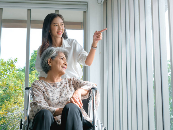Щаслива азіатська літня жінка, мати або дідусь з бабусею на інвалідному візку, які доглядають за доглядачем, усміхаються молодій жінці, дочці або внучці, дивляться у вікно, підтримуючи вдома, старшій охороні здоров'я. - Фото, зображення