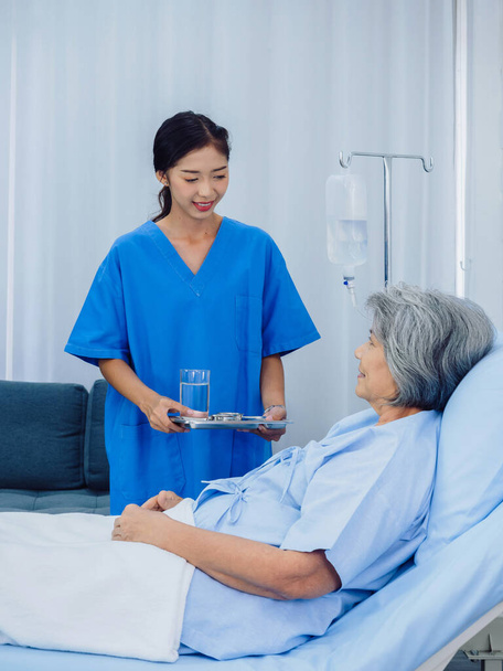 Freundlich lächelnde junge asiatische Krankenschwester im blauen Peeling, die Tablett mit Tabletten für ältere Patienten hält, die im Krankenhauszimmer auf dem Bett liegen und Medikamente oder Vitaminpräparate einnehmen.. - Foto, Bild