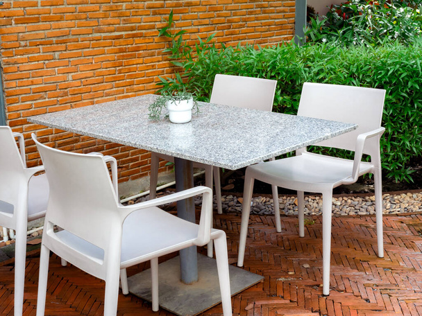 Cuatro sillas vacías de plástico blanco con mesa de terrazo con maceta pequeña en el jardín al aire libre cerca del suelo de ladrillo y el edificio de la pared. Conjunto de mesa relajante en jardín. - Foto, imagen