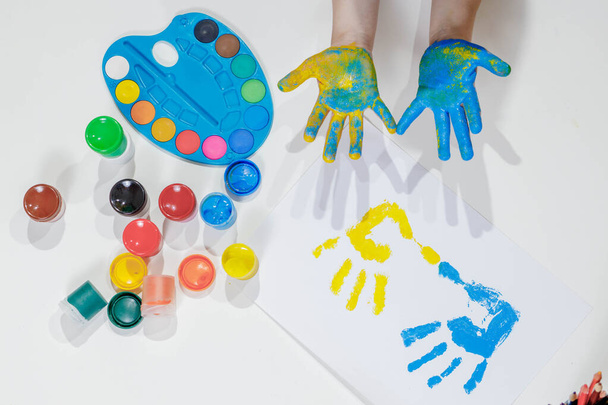 Children's handprints. Blue and yellow palm prints on a white sheet of paper, children's hands in paint, a palette of colorful paints, gouache. Top view of the table. Children's creativity. - Foto, Imagem