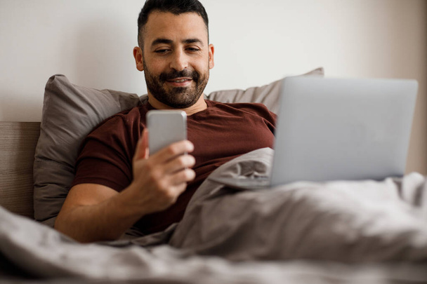 Freiberufler im Schlafanzug mit Laptop und Telefon, während er zu Hause im modernen Schlafzimmer liegt. Hochwertiges Foto - Foto, Bild