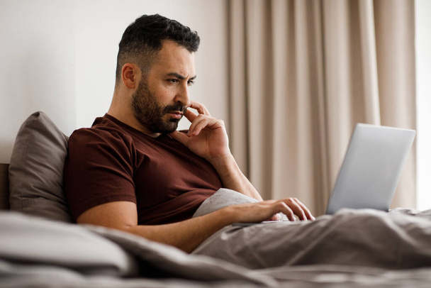 Lachende freelancer in home kleding werken aan moderne laptop op comfortabel bed thuis, zijaanzicht. Hoge kwaliteit foto - Foto, afbeelding