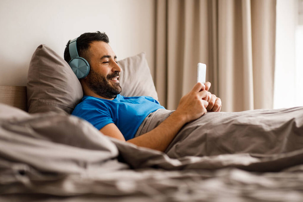 Счастливый ближневосточный парень в беспроводных наушниках отдыхает в спальне с мобильным телефоном в чате с друзьями или слушает музыку. Высокое качество фото - Фото, изображение