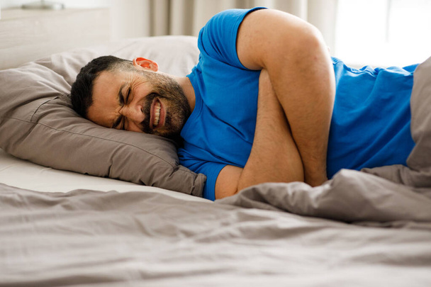 Widok z boku młodego brodatego mężczyzny w piżamie leżącego w łóżku z zamkniętymi oczami i cierpiącego na ból brzucha. Wysokiej jakości zdjęcie - Zdjęcie, obraz