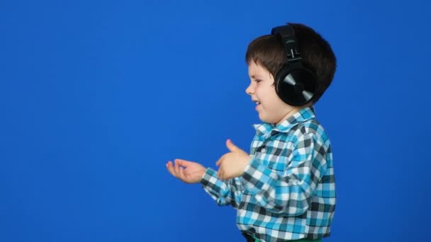 Een 5-jarige jongen met een koptelefoon klapt vrolijk en droevig met zijn handen op een blauwe achtergrond - Video