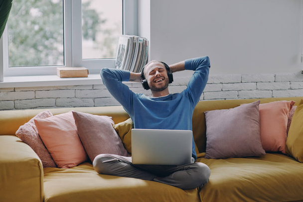Relaxed man in headphones holding hand behind head and smiling while sitting on the couch - Photo, Image