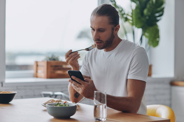 A fiatalember élvezi az ebédet és okos telefont használ, miközben a konyhában ül. - Fotó, kép