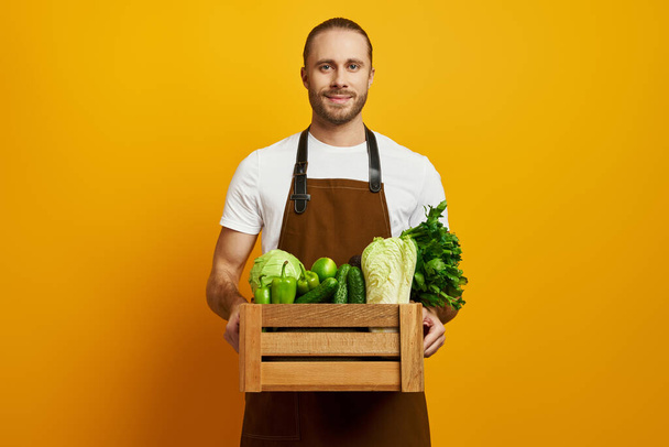 Confident young man in apron carrying wooden crate with fresh veggies against yellow background - Foto, Imagem