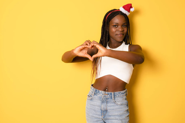 Retrato de hermosa mujer afroamericana modelo con sombrero de santa haciendo corazón con los dedos aislados sobre fondo amarillo. - Foto, imagen