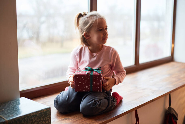 Little caucasian girl unpacks gift boxes at home near window. Happy family christmas concept - 写真・画像