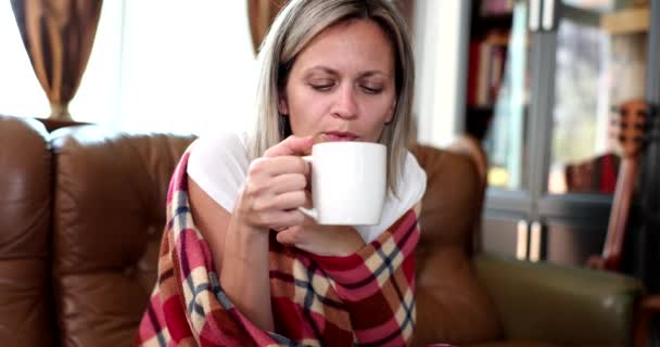 Mulher bonita é enrolado em cobertor quente com xícara quente no sofá em casa. Calafrios febre e bebida quente - Filmagem, Vídeo