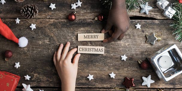 Manos de niños caucásicos y negros haciendo un alegre letrero de Navidad escrito en clavijas de madera colocándolas en un escritorio rústico de madera lleno de adornos y decoración navideña.. - Foto, Imagen